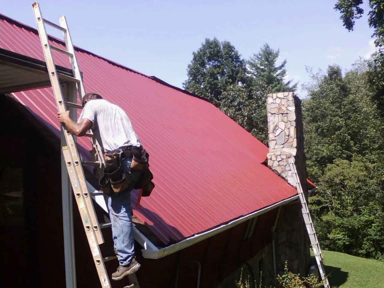 Roofer on a ladder beside a red metal roof and stone chimney — Soffit & Fascia Repair by Roofing Brothers in Scarville